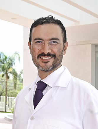 Es la foto de perfil de un cirujano plástico en León de Hospital Angeles León. Utiliza una bata blanca con una camisa. Tiene una corbata negra, lentes y está sonriendo.