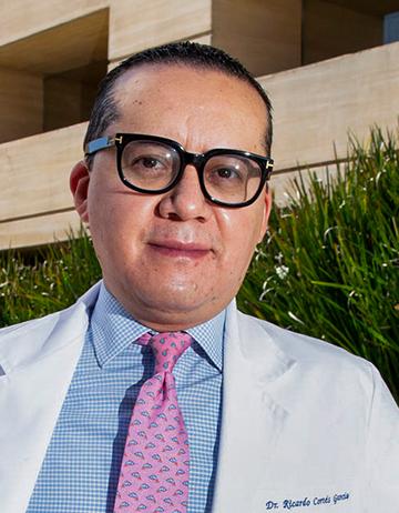 Es la foto de perfil de un cirujano pediátrico en Ciudad de México de Hospital Angeles. Utiliza una bata blanca, camisa azul con cuadros blancos y una corbata rosa.
