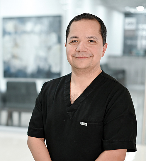 Es la foto de perfil de un especialista en reproducción asistida y en ginecología y ibstetricia en Ciudad de México de Hospital Angeles. Viste una una camisa color lila y una corbata de color verde claro con figuras blancas y trae sobre la ropa una bata blanca. Es de tez morena, está sonriendo suavemente a la cámara. 